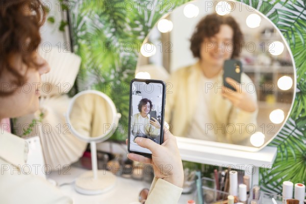 A person takes a selfie using a smartphone in front of a round, illuminated vanity mirror The background features a vibrant, leafy wallpaper The scene captures a candid, modern moment