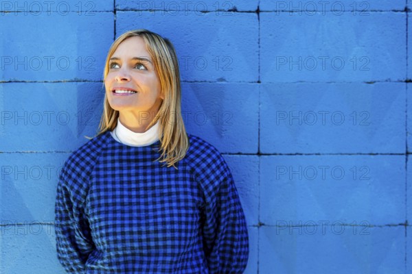 A blonde woman in a plaid shirt gazes upward, standing against a vivid blue background