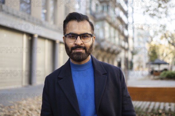 Indian businessman in a stylish winter outfit with a blue sweater and dark coat, standing outdoors, looking at the camera with a gentle smile, amidst an urban setting