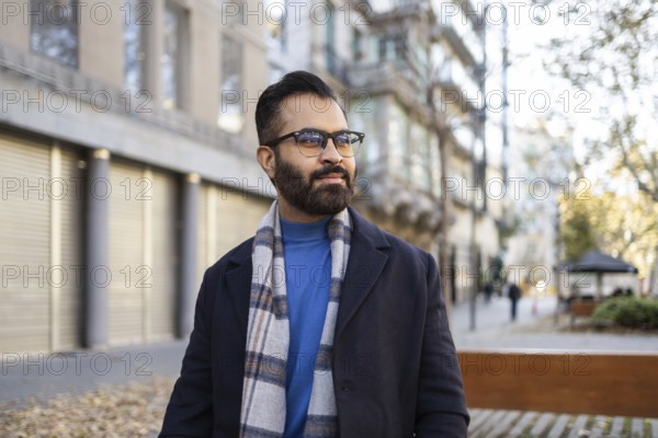 An Indian man dressed in a stylish winter coat and scarf, standing confidently in an urban, outdoor environment. He looks away thoughtfully, sporting a beard and glasses