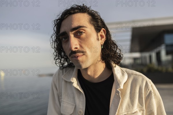 A man with curly hair and a mustache looks confidently at the camera He wears a casual light jacket over a black shirt, with a blurred modern building background