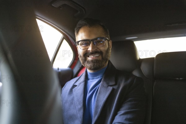 Smiling Indian businessman wearing glasses and winter attire seated in a car, looking directly at the camera