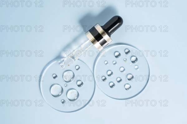 A pipette dropper with a skincare serum rests on a smooth, light blue surface Surrounding the pipette are clear serum drops adding a minimalistic and fresh aesthetic