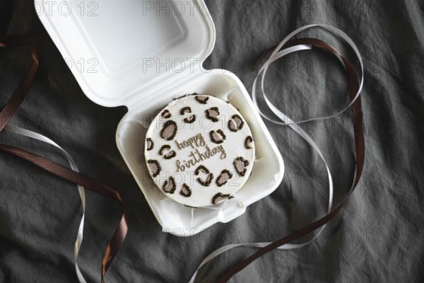 Top view of a stylish bento birthday cake with a Happy Birthday message, decorated with animal print shapes. The cake is nestled in a white takeout box, complemented by elegant ribbons on a textured grey background