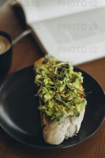 A close-up of avocado toast topped with greens and seeds, served on a black plate. A cup of coffee and an open book create a cozy atmosphere on a wooden table