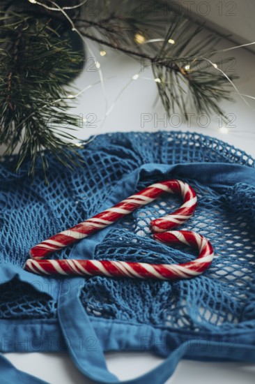 A blue string bag overflowing with red and white striped candy canes, placed alongside a pine branch adorned with twinkling lights, creates a cozy wintertime ambiance