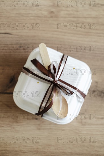 Top view of an elegantly packaged bento birthday cake, tied with a brown satin ribbon and wooden spoon, set on a rustic wooden background