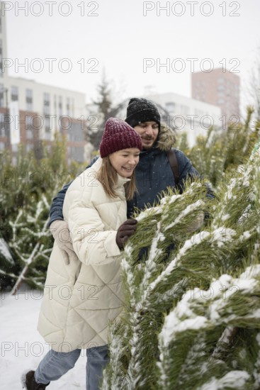 A couple enjoys picking out a Christmas fir at an outdoor market during a snowy day They are dressed warmly in winter coats and hats, surrounded by festive greenery