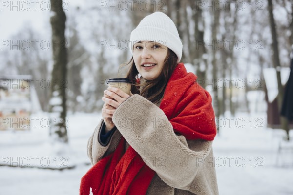 A young woman in winter clothing holds a coffee cup while wrapped in a cozy, red scarf, in a snowy landscape