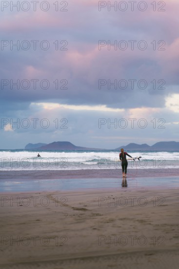 Surfers in a wetsuit approach the waves under a dramatic, colorful sky on a Lanzarote beach. The morning light and distant mountains create a serene atmosphere
