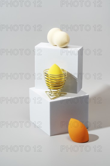Three cosmetic sponges in different colors rest on minimalist white geometric stands against a soft background, highlighting their vibrant hues and smooth texture