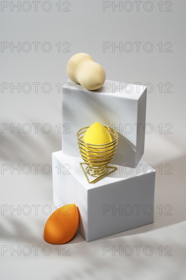 Vibrant cosmetic sponges in orange and yellow, elegantly arranged on minimalist white platforms with golden holders, softly lit against a neutral background