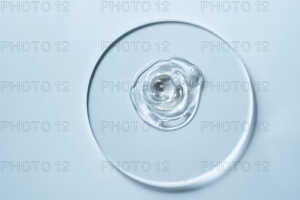 A clear gel with small golden specks on a round glass plate against a light blue background The minimalistic composition highlights texture and transparency
