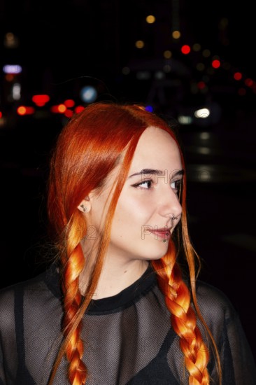 A woman with striking red braids and facial piercings stands against a backdrop of blurred city lights at night. She wears a sheer black top, capturing urban style and individuality