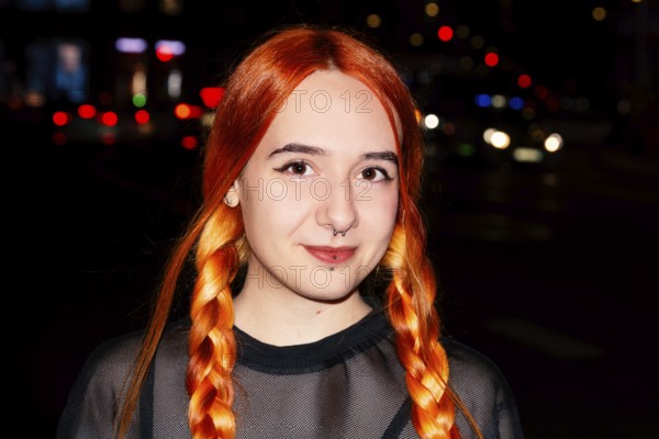 A young woman with bright orange hair styled in braids is smiling against a blurred urban night background with colorful lights. She wears a black mesh top and septum piercing