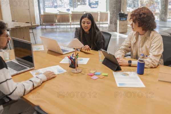 A diverse group of coworkers engaged in a collaborative meeting around a large wooden table in a modern office setting, using laptops and tablets for teamwork and discussion