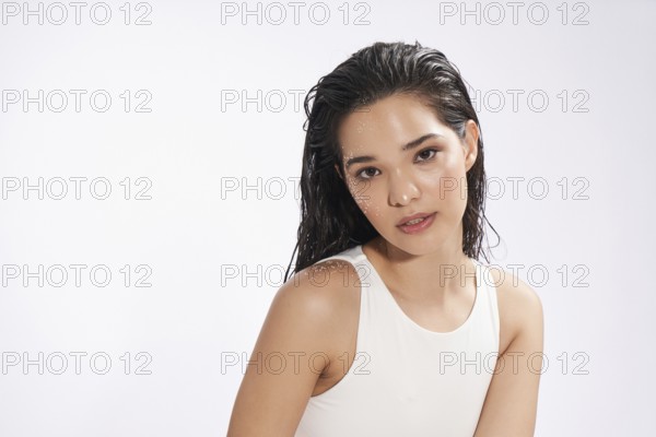 A young woman with wet hair and water droplets on her skin poses in a studio, looking directly at the camera. She wears a simple white bodysuit, exuding a natural and fresh appearance