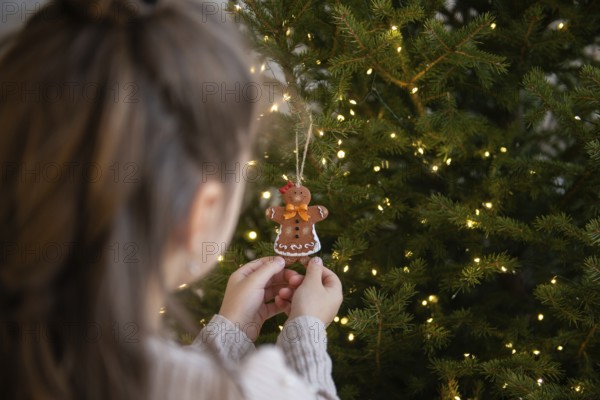 Back view of crop unrecognizable young girl, carefully hangs a gingerbread man ornament on a brightly lit Christmas tree. The twinkling lights add a warm, festive glow to the scene