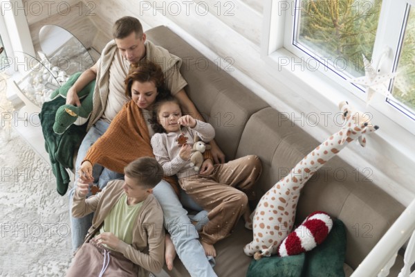 Top view of a festive setting with a winter view and holiday deco. A joyful family celebrates Christmas together on a couch. A couple relax with two kids, an older boy and a younger girl. Both children are engrossed with toys