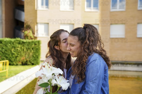 A young LGBTQ+ women couple, sharing a tender moment outside, with one women kissing the other on her cheek while holding white lilies, in front of a residential building