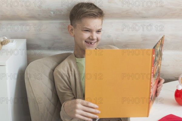 A young boy with short, blond hair enjoys reading a vibrant orange book, his engaging smile reflecting a moment of joy. Clad in a beige jacket over a green tee, he represents a cozy, scholarly aesthetic, seated in a softly lit room