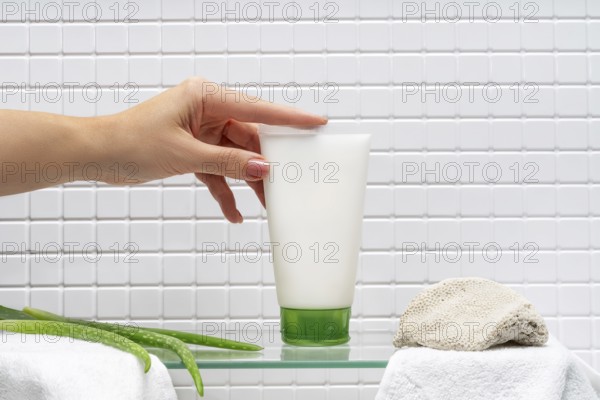 Cropped unrecognizable woman holding a tube of body care cream beside aloe gel in a bathroom, with a white towel and loofah sponge, on a glass shelf against a tiled wall