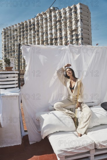 A woman lounges on a rooftop with white linens and cushions, basking in the summer sun The backdrop of a modern building adds an urban contrast to the relaxed scene