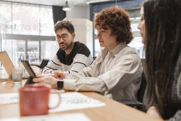 Three coworkers discuss projects around a wooden table in a bright, modern office They share ideas while using laptops and smartphones, fostering creativity and teamwork