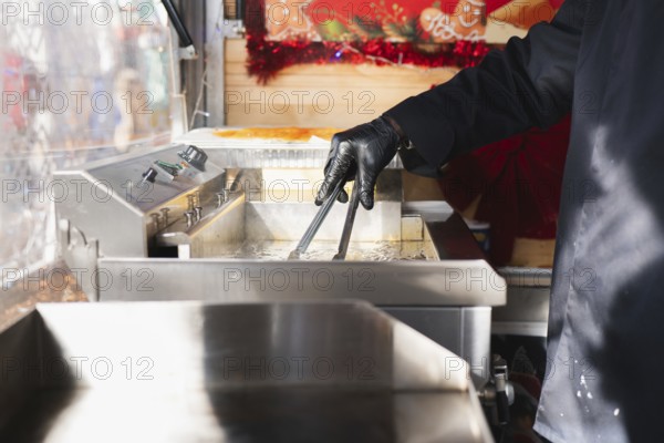 A hand in a black glove uses tongs to fry spiral tornado potatoes in an outdoor food stall setting. The stainless steel fryer and festive decor create a vibrant market atmosphere