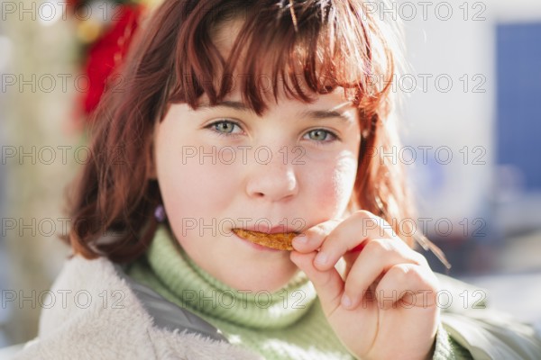A young girl with red hair and green eyes enjoys a crunchy snack outdoors. The scene captures the play of sunlight on her face, evoking a sense of childhood joy and leisure