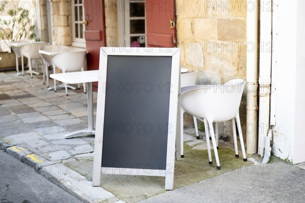 A blank chalkboard standing outside a cozy cafe with white chairs and tables, against a stone wall. The scene invites customization for events, menus, or promotions