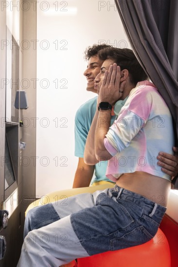 A joyful gay couple sits closely in a booth, sharing laughter and intimate expressions. They are dressed in casual, colorful clothing, creating a warm and affectionate atmosphere