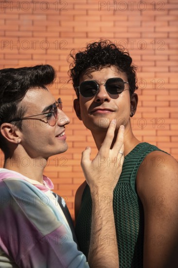 A gay couple shares a tender moment, smiling in the sunlight. They wear sunglasses and casual outfits, set against a vibrant brick wall, highlighting their joyful relationship