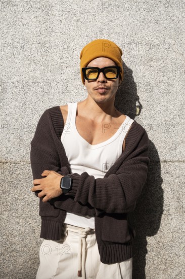 A stylish Colombian gay man leans against a concrete wall, wearing a yellow beanie, sunglasses, white tank top, and black cardigan, exuding confidence in an urban setting