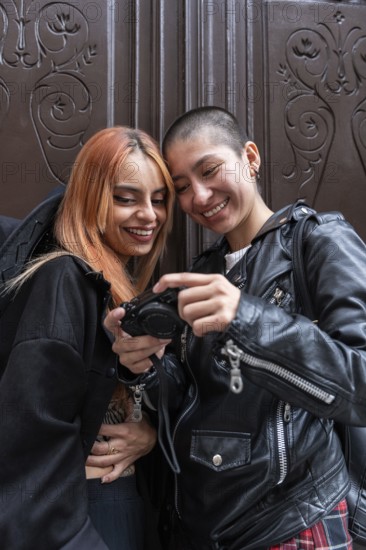 Two joyful lesbian women share a moment, looking at photos on a camera while leaning against a decorative wooden door. They smile together, enjoying each other's company