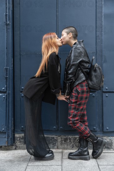 A stylish lesbian couple shares a tender kiss in front of a blue urban background. They exude confidence and love, each dressed in unique, bold fashion