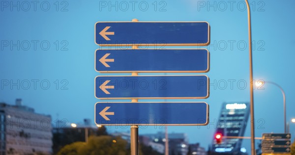 A street sign with multiple arrows pointing to the left, set against a cityscape. The scene captures the essence of urban commuting and navigation at dusk's blue hour