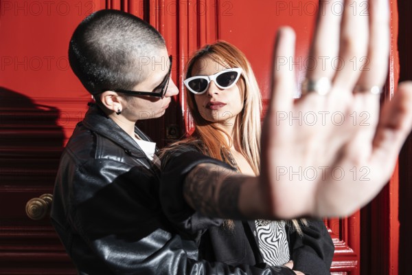 A fashionable lesbian couple stands confidently against a bold red door. One person playfully raises a hand, while both wear sunglasses, showcasing a modern, urban vibe