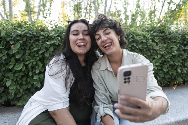 A cheerful lesbian couple takes a selfie with a smartphone in a garden. They smile brightly, enjoying a sunny day. Their happiness and love are captured in this candid moment