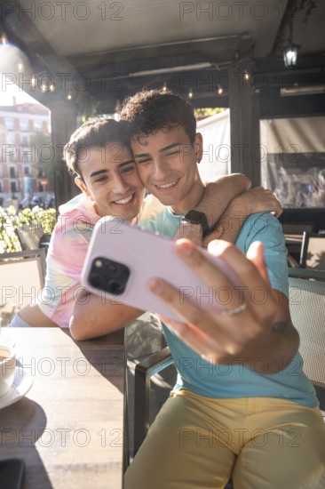 A joyful moment captured as a gay couple take a selfie at an outdoor cafe. They are embracing warmly, surrounded by light and lively atmosphere
