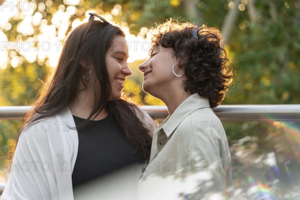A lesbian couple shares a tender moment, smiling warmly at each other in the soft glow of sunset, symbolizing love and connection against a backdrop of vibrant greenery
