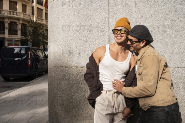A smiling Colombian couple shares a tender moment on a city street. Dressed casually and stylishly, they stand against a textured wall, enjoying the sunny day together