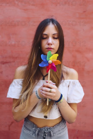 A teenage girl with long hair holds a vibrant pinwheel against a coral backdrop. She appears calm and reflective, enjoying a sunny day. Her casual attire adds to the relaxed mood