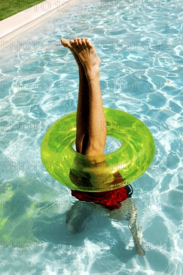 A person playfully balances upside down in a swimming pool, legs pointing skyward through a bright green float ring, capturing a fun, carefree summer vibe under the sun