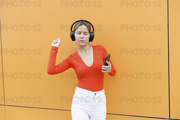 Woman in a red top using a smartphone and wearing headphones, standing against an orange wall in Warsaw She appears to be enjoying music or a podcast