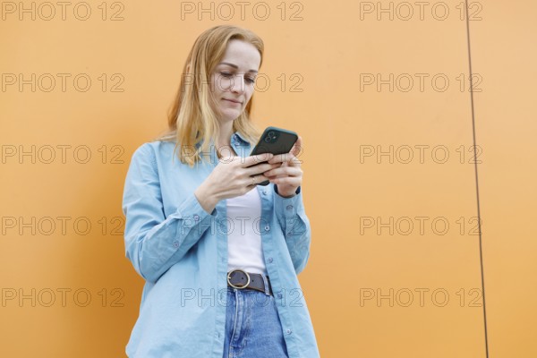 A woman stands against an orange wall in Warsaw, intently using her smartphone She wears a blue shirt and jeans, focusing on her screen, embodying modern life in an urban setting