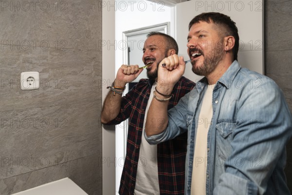 Two gay men in casual shirts joyfully brush their teeth together in a modern bathroom, emphasizing a harmonious routine. The scene captures everyday happiness and companionship