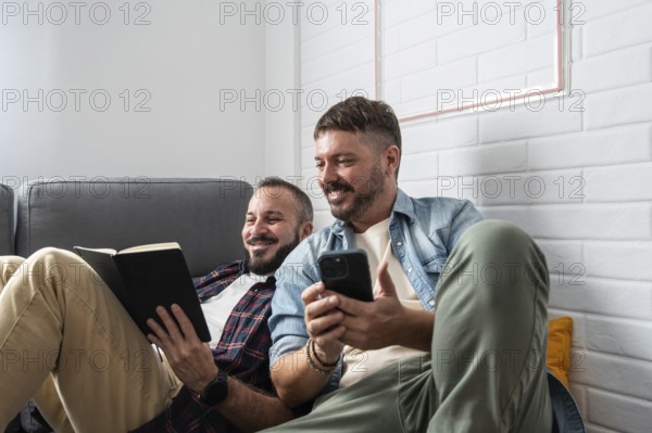 A joyful gay couple relaxes on a cozy sofa, one reading a book and the other holding a smartphone. They are enjoying a peaceful moment together in their modern living room