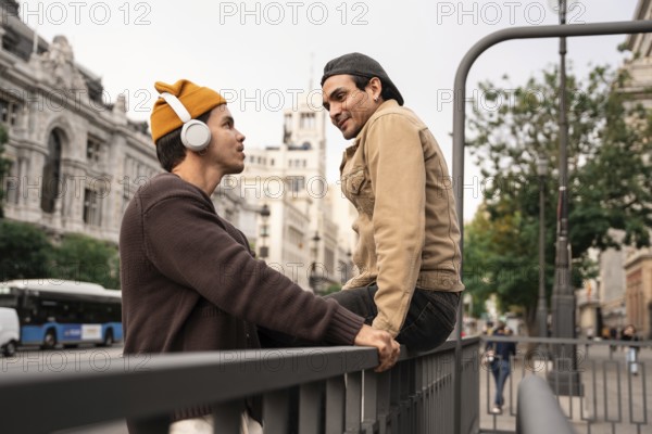 A Colombian couple enjoys a moment together in Madrid city, one listening to music with headphones, the other sitting on a railing with vibrant expressions