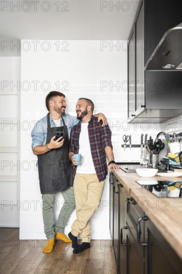 A gay couple standing close in a modern kitchen, sharing a happy moment. One wears an apron and holds a phone, the other holds a mug, both smiling warmly